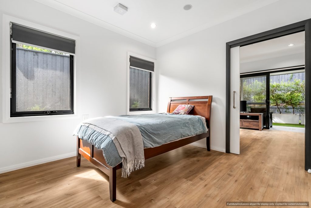 A bright bedroom in Inner West Sydney’s Concord, featuring a wooden bed, blue bedding, two windows with blinds, and a sliding door opening to another room with large windows and greenery outside. 10E.