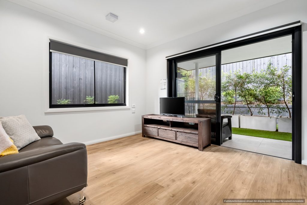 Modern living room in Concord, Inner West Sydney with wooden flooring, a brown sofa, TV on a wooden stand, large window, and sliding glass doors opening to a covered patio with greenery outside.
