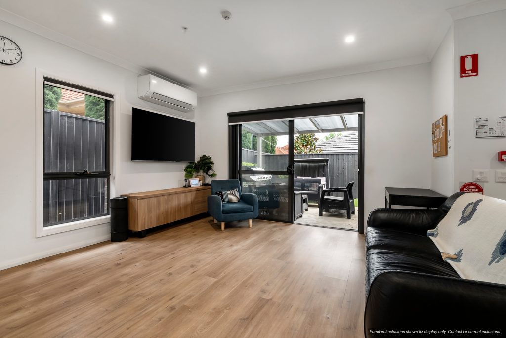 Modern living room in Concord, Inner West Sydney, featuring a wooden floor, black sofa, blue armchair, wall-mounted TV, air conditioner, and sliding glass door opening to a covered patio.