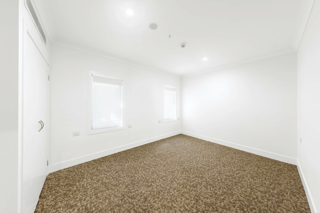 A vacant, well-lit room with white walls, two windows with blinds, beige carpet flooring, and a closed white door on the left side in the heart of Maroubra in Sydney’s Eastern Suburbs.