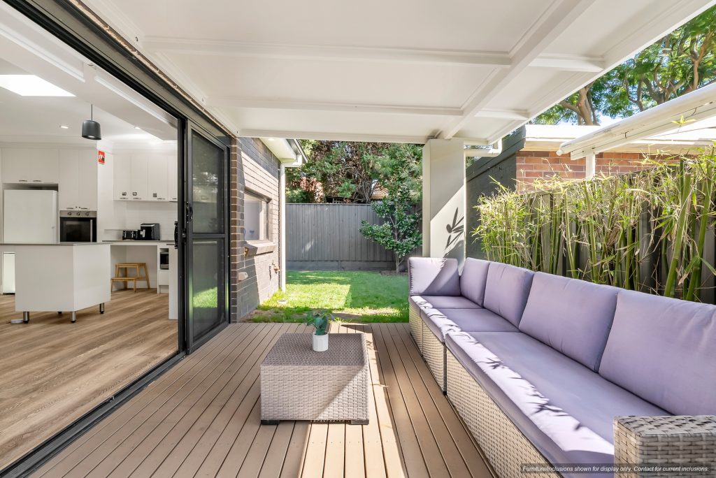 Modern covered patio in Sydney’s Eastern Suburbs with a long cushioned outdoor sofa, coffee table, wooden deck, and sliding glass doors opening to a kitchen and small fenced backyard.