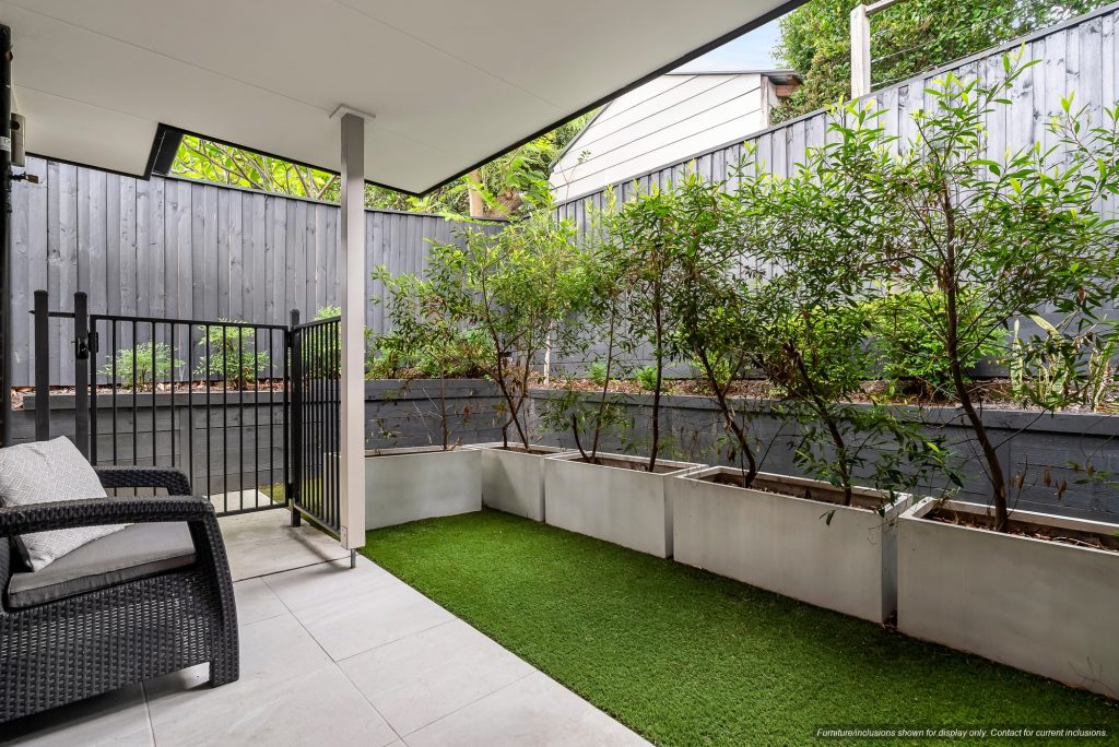 A small, covered patio with tiled flooring, outdoor seating, and a fenced area leading to a garden bed with shrubs and artificial grass—perfect for enjoying Concord’s relaxed lifestyle in 10E, Inner West Sydney.