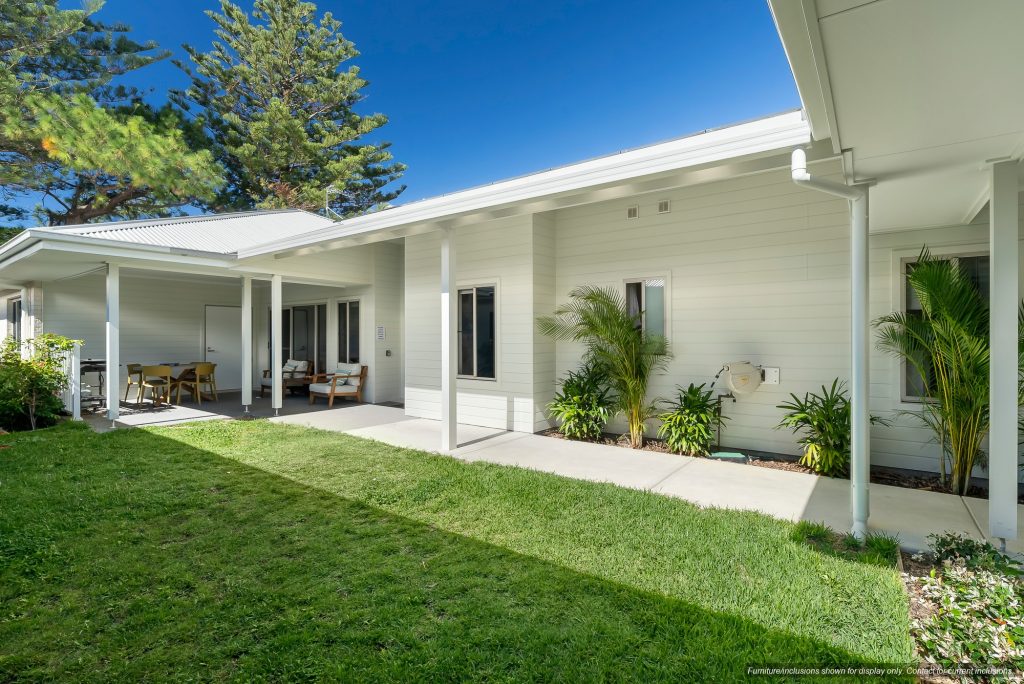 A modern single-story house in Narraweena, Northern Beaches, Sydney, featuring white siding, a covered patio with outdoor seating, and a well-maintained green lawn under a clear blue sky.