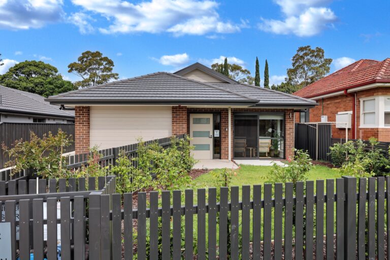 Single-story brick house with a gray tiled roof, a front lawn, and a black picket fence located in Concord, Inner West Sydney. A paved path leads to the front door. Trees and blue sky with clouds are in the background.