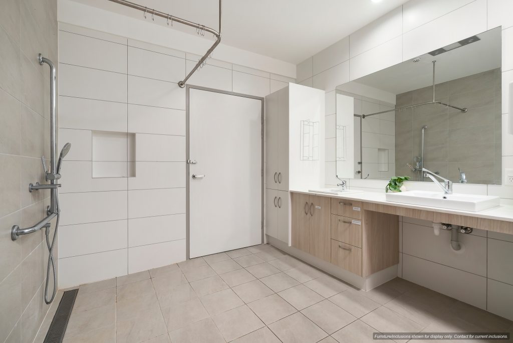 Spacious, accessible bathroom in Narraweena, Northern Beaches, Sydney, featuring a walk-in shower, grab bars, handheld showerhead, large mirror, and wooden cabinetry beneath a wide sink.