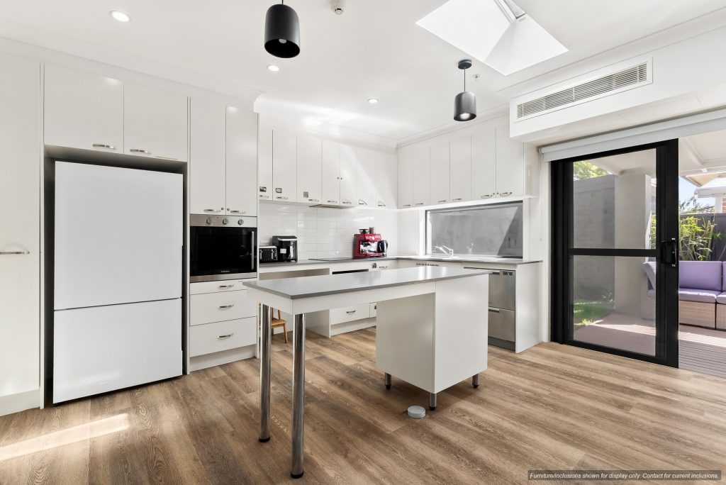 Modern kitchen in Maroubra with white cabinets, stainless steel appliances, central island on wheels, wood flooring, large windows, and a sliding glass door leading to an outdoor patio—perfect for enjoying Sydney’s Eastern Suburbs lifestyle.
