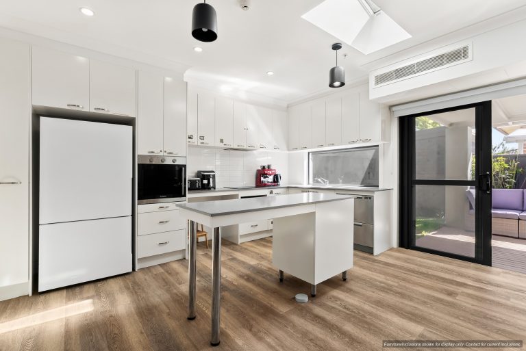 Modern white kitchen in Maroubra with stainless steel appliances, central island on wheels, wood flooring, and sliding glass door leading to an outdoor patio in Sydney’s Eastern Suburbs.