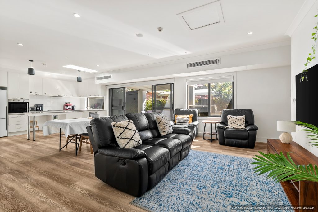 Contemporary open-plan living room and kitchen in Maroubra, Sydney, featuring black leather sofas, wooden flooring, white cabinetry, dining table, large windows, and a blue rug—perfect for relaxed Eastern Suburbs living.