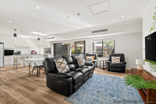 Contemporary open-plan living room and kitchen in Maroubra, Sydney, featuring black leather sofas, wooden flooring, white cabinetry, dining table, large windows, and a blue rug—perfect for relaxed Eastern Suburbs living.
