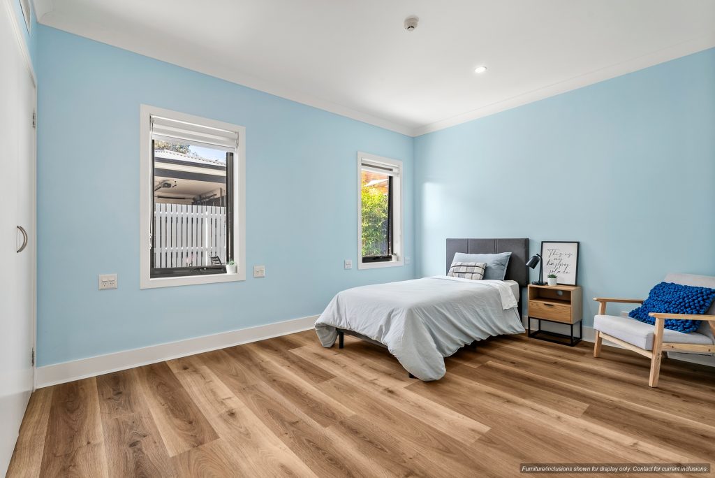 Minimalist bedroom in Maroubra with light blue walls, single bed, side table, armchair, and two windows for natural light. Wood flooring and simple decor enhance the modern look of this Eastern Suburbs Sydney home.