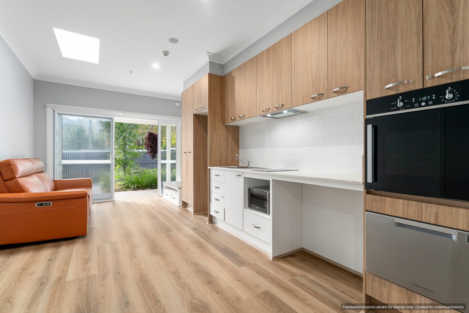 Modern studio apartment in Pagewood with wood flooring, built-in kitchen cabinets, white backsplash, brown reclining chair, and glass doors opening to a small patio with garden view in Inner South Sydney.
