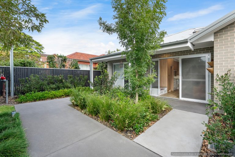 A modern single-story house in Pagewood with a light brick exterior, large windows, and a glass door, surrounded by a landscaped garden and concrete pathways in sought-after Inner South Sydney.