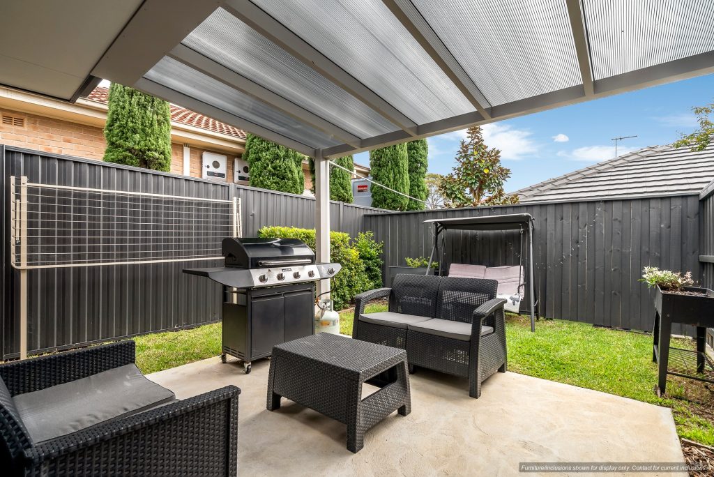 Covered patio in Concord, Inner West Sydney, featuring black wicker furniture, a barbecue grill, a swing bench, and a clothesline, all enclosed by a black fence with green bushes and trees in the background.