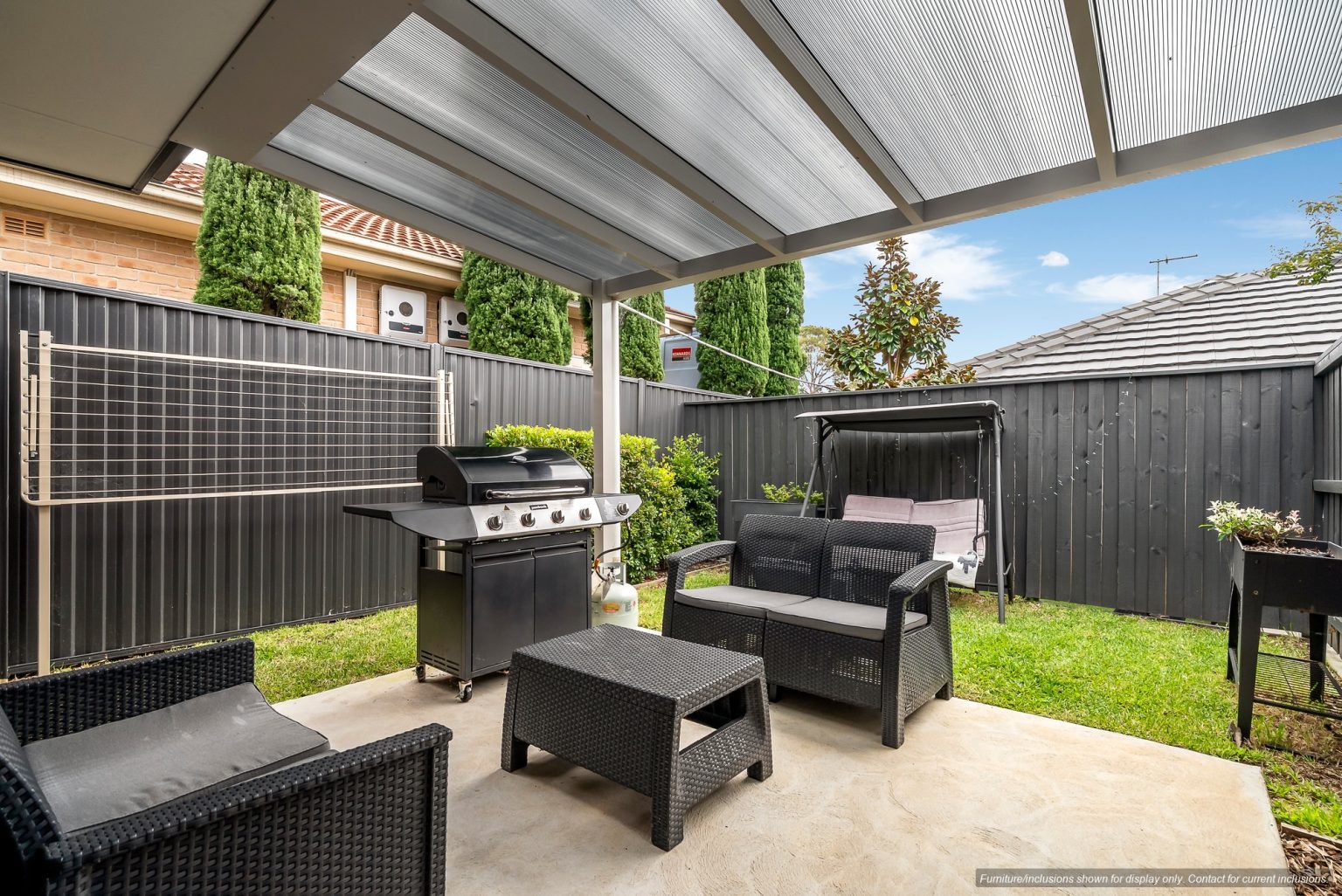 Covered patio in Concord, Inner West Sydney, featuring black wicker furniture, a barbecue grill, a swing bench, and a clothesline, all enclosed by a black fence with green bushes and trees in the background.