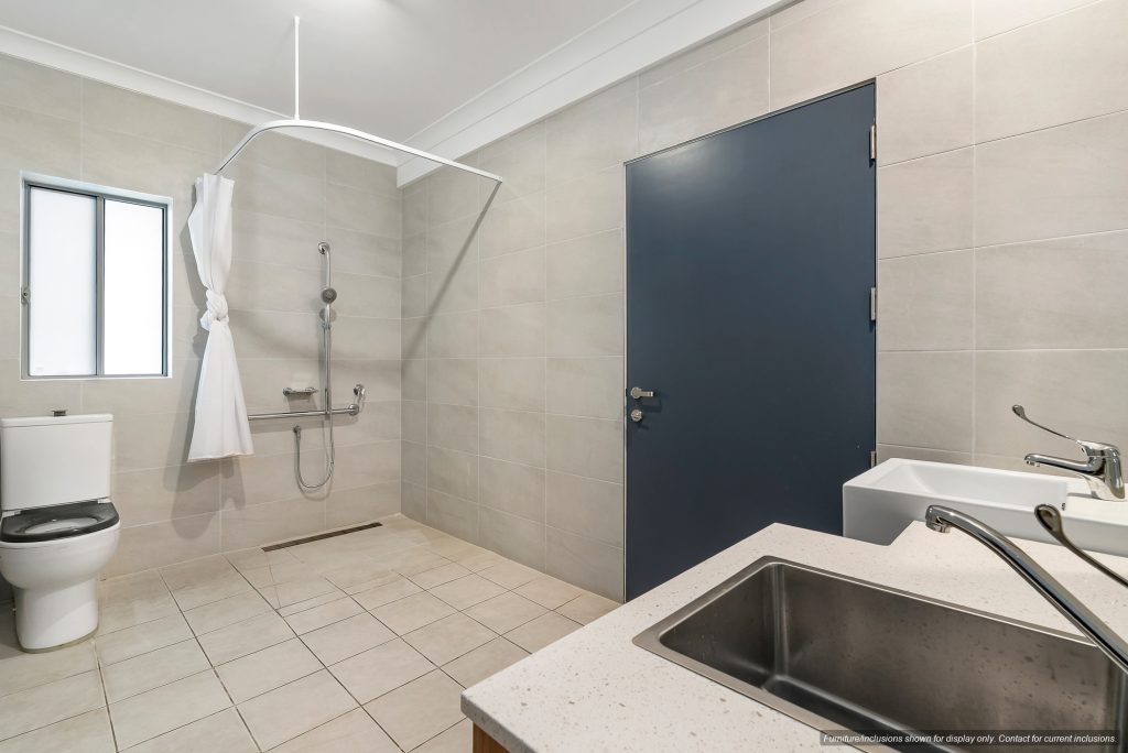 Accessible 9B bathroom with a tiled floor, open shower area with grab bars, toilet, sink, and a blue door. Located in Inner South Sydney’s Pagewood. A white curtain is tied to the shower rail.