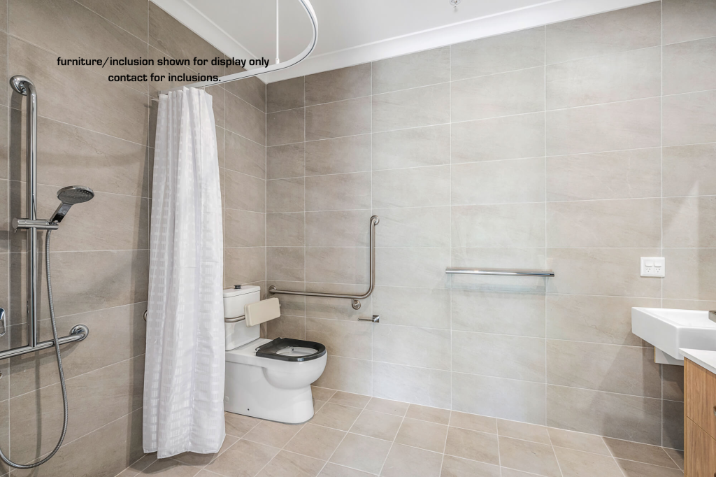 Accessible bathroom at D Wamberal with beige tiles, walk-in shower, wall-mounted grab bars, toilet, white shower curtain, and sink. Text on wall notes display items may not be included.