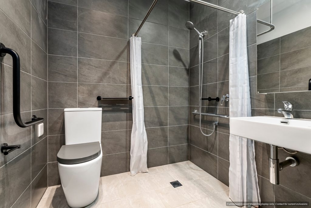 Modern accessible bathroom in Concord, Inner West Sydney, with gray tiled walls, a white toilet, wall-mounted sink, walk-in shower, grab bars, and white shower curtains.
