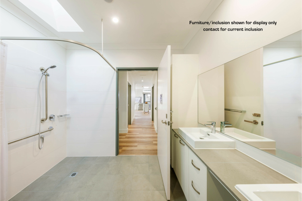 Spacious, accessible bathroom with a walk-in shower, double sinks, and a large mirror in sought-after Maroubra. Open door reveals hallway with wooden flooring. Text about furniture inclusion is displayed on the wall in this Eastern Suburbs Sydney home.