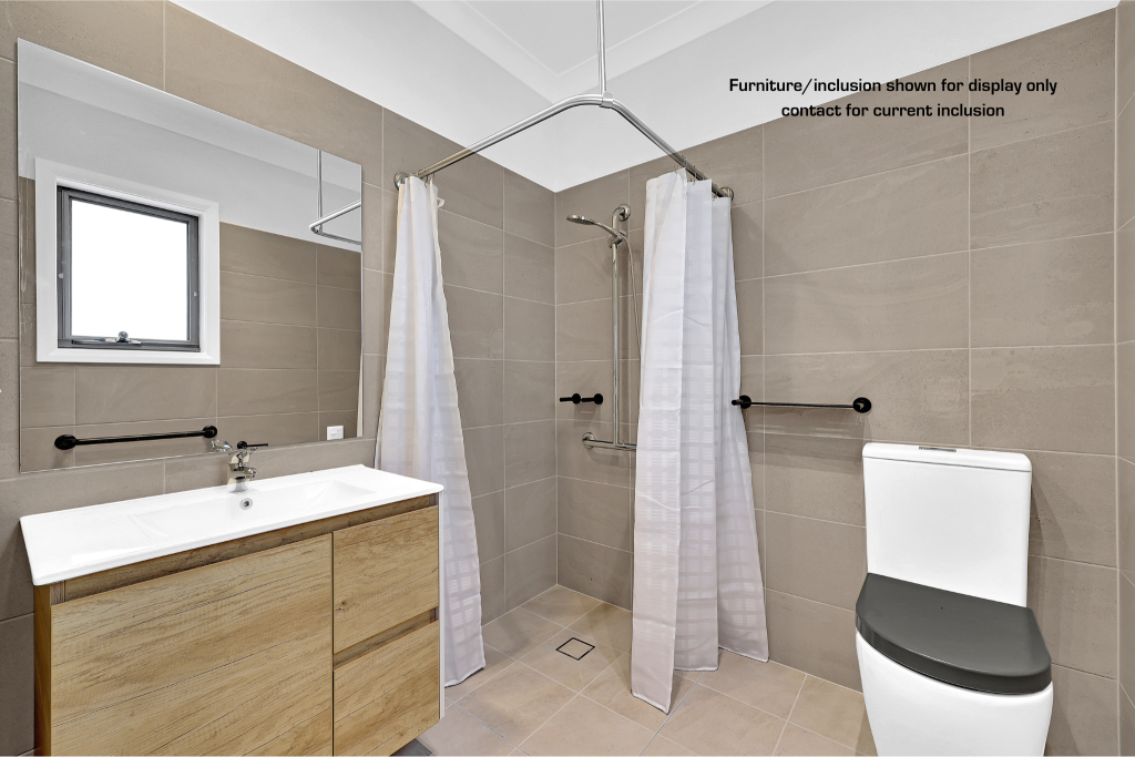 Modern accessible bathroom in Concord, Inner West Sydney, with beige tiles, a white toilet, wooden vanity, wall-mounted sinks, grab bars, and a walk-in shower with white curtains. Text on wall provides display disclaimer.