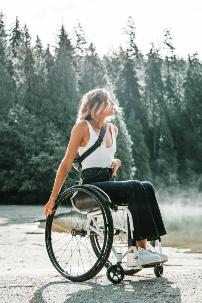 A woman in a wheelchair outdoors, enjoying sunlight with a forest backdrop. she has blonde hair and wears a white top and black pants.
