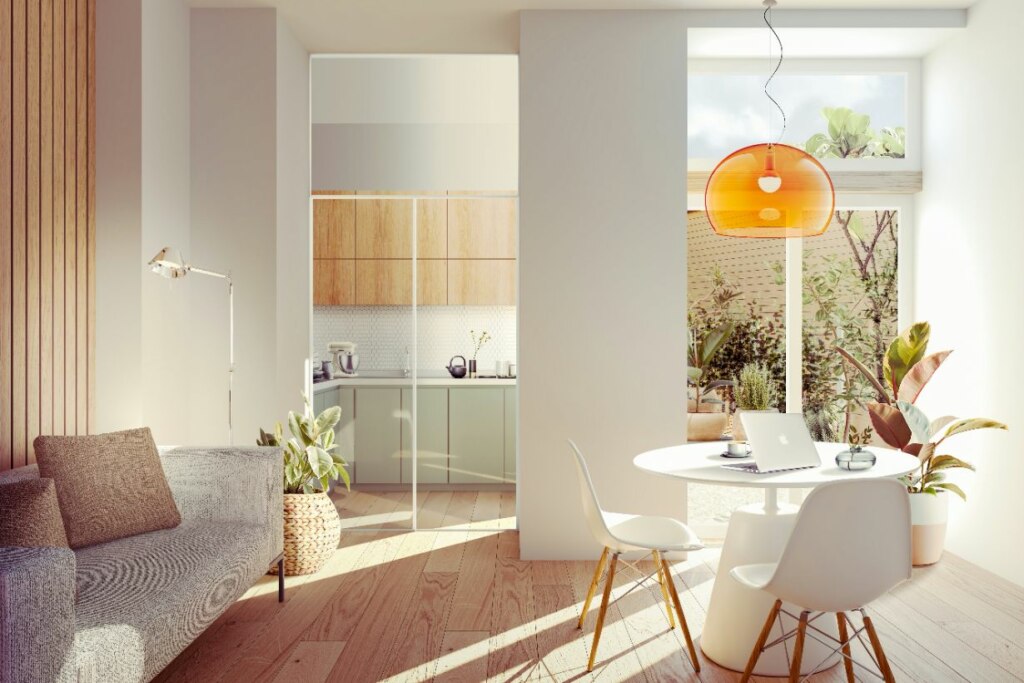 Modern sunlit living room with kitchen in the background, showcasing a sofa, a white dining table with chairs, laptop, and hanging orange lamp, complemented by indoor plants.