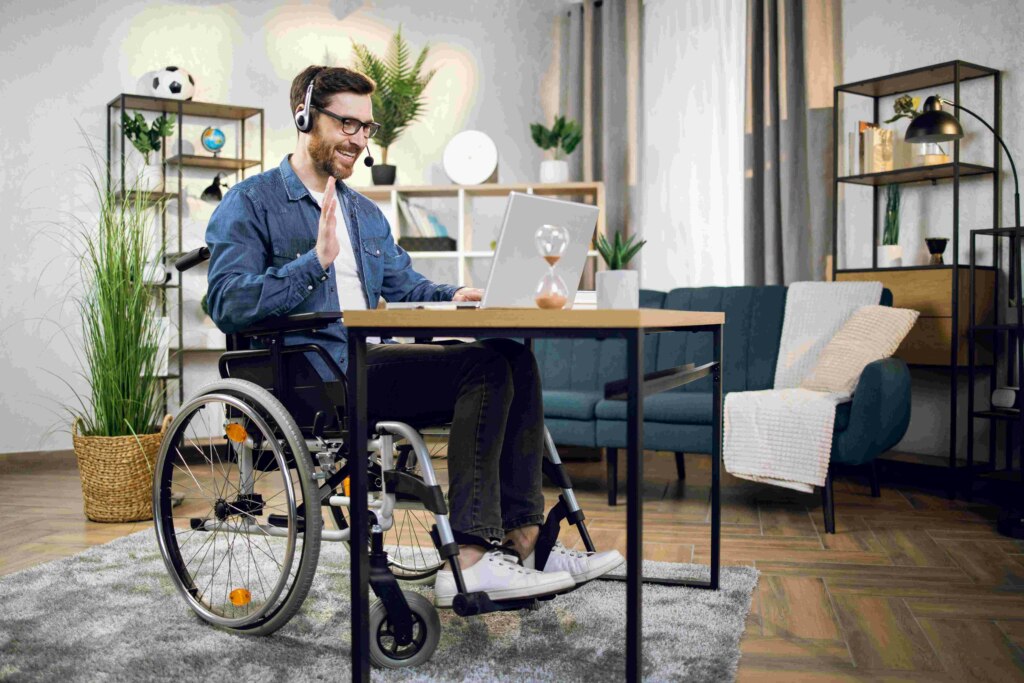 Man in a wheelchair at a desk with a laptop, smiling and clapping during an NDIS video call.