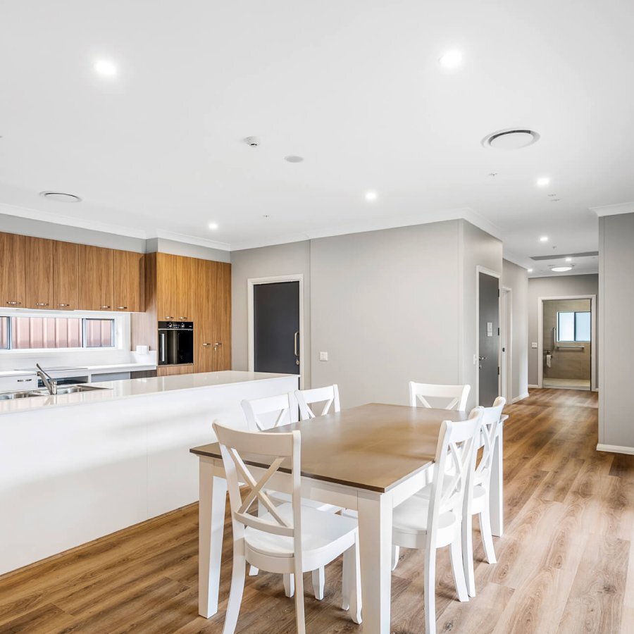Modern kitchen and dining area with wooden cabinets, white countertops, a wooden dining table with white chairs, and hardwood flooring. Doorways lead to other parts of the house.