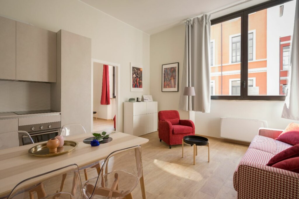 A bright, modern NDIS apartment living room with a dining area, red armchair, white sofa, and large windows overlooking the city.
