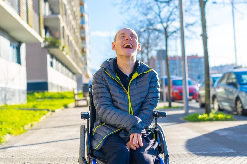 A person in a wheelchair smiles and laughs outdoors on a sunny day, with buildings, trees, and parked cars in the background.