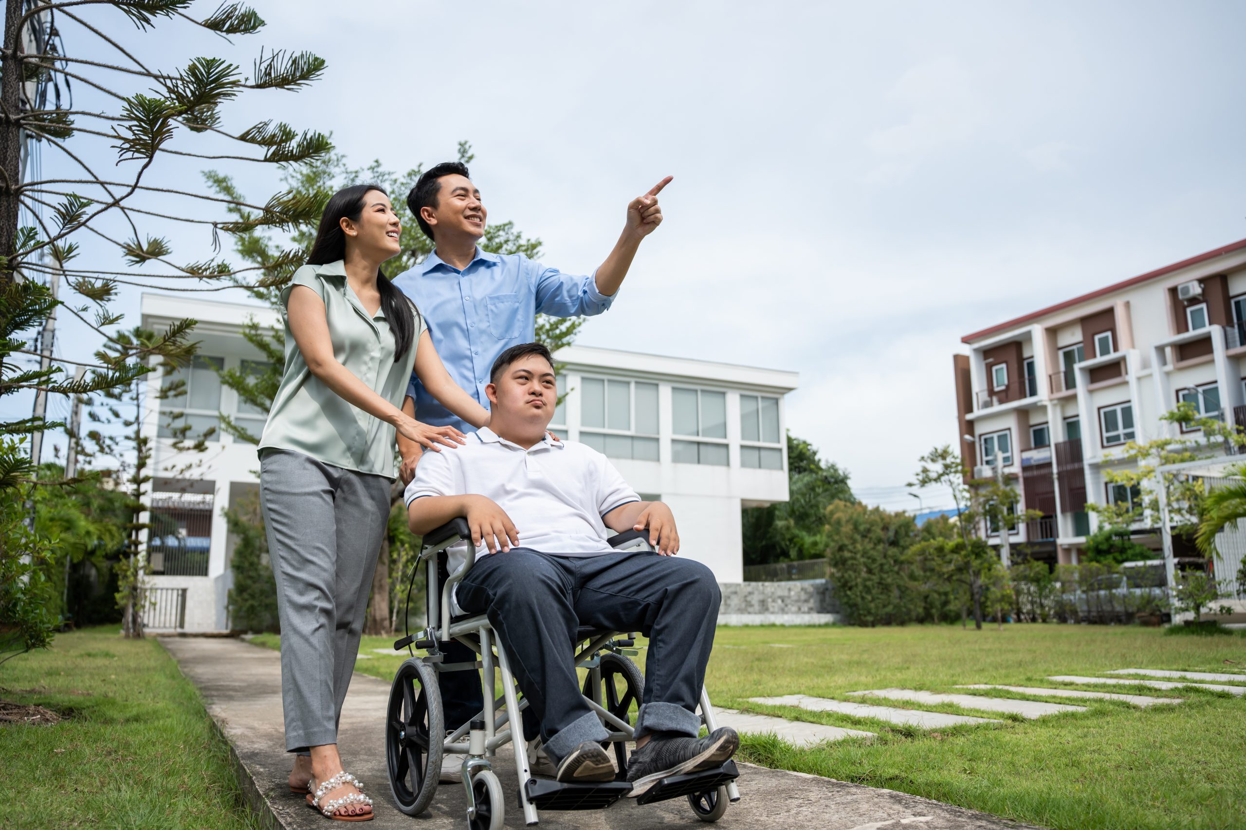 Three people outdoors near modern buildings; one person sits in a wheelchair while the other two stand beside and behind, with one person pointing ahead.