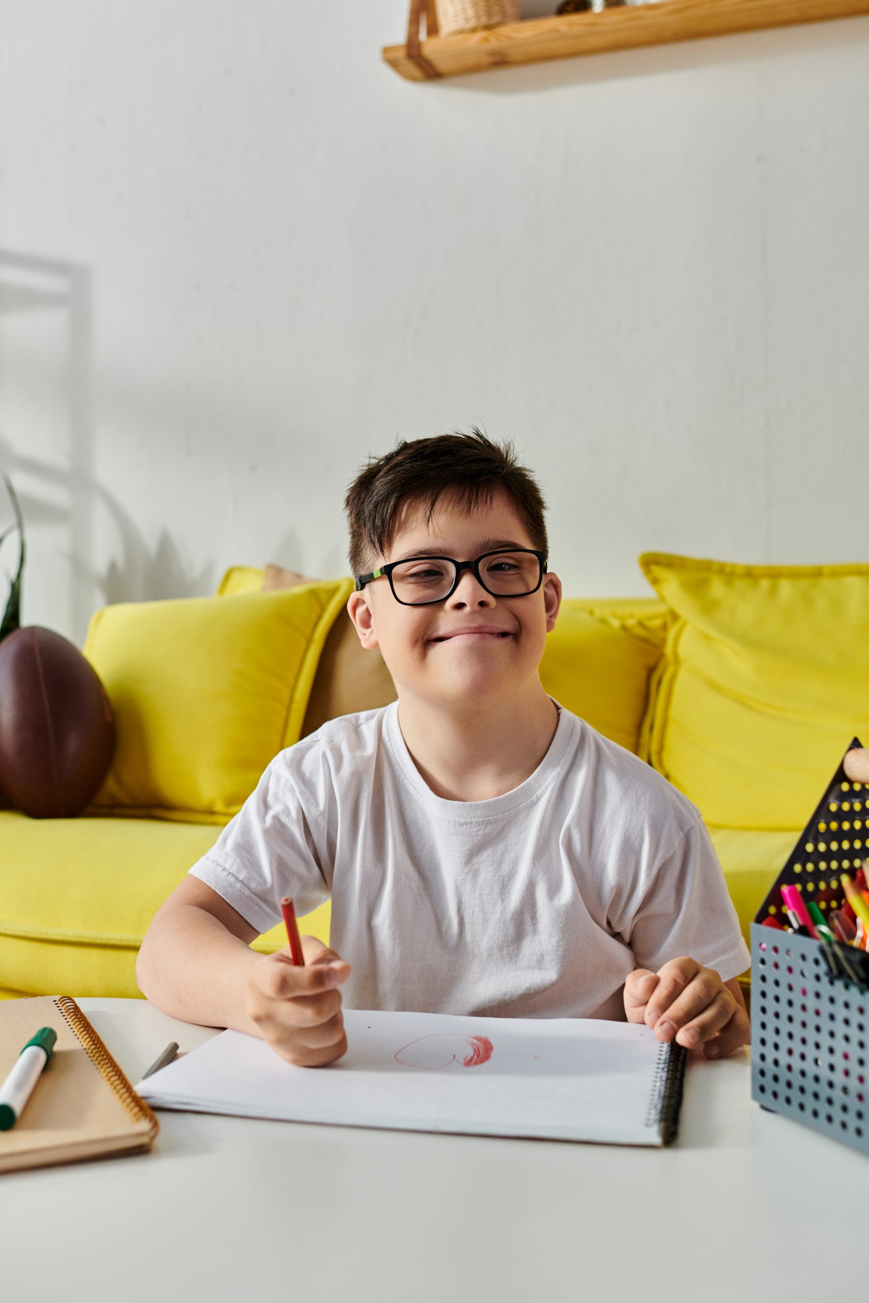 A boy with Down syndrome wearing glasses and a white t-shirt sits at a table, drawing with colored pencils in a sketchbook, with a yellow couch in the background.