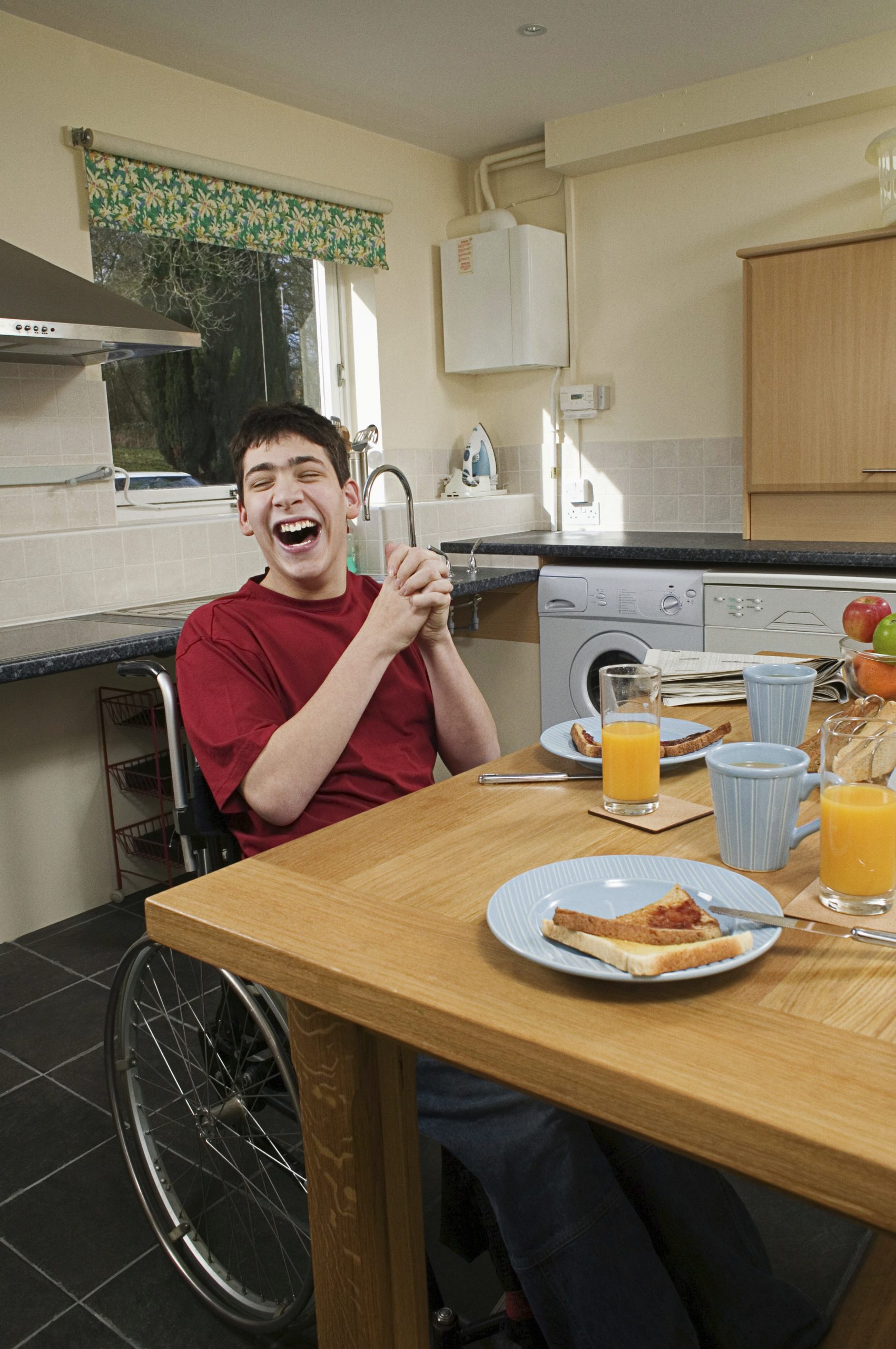A young person in a wheelchair smiles at a kitchen table set with plates of toast, cups, and glasses of orange juice.