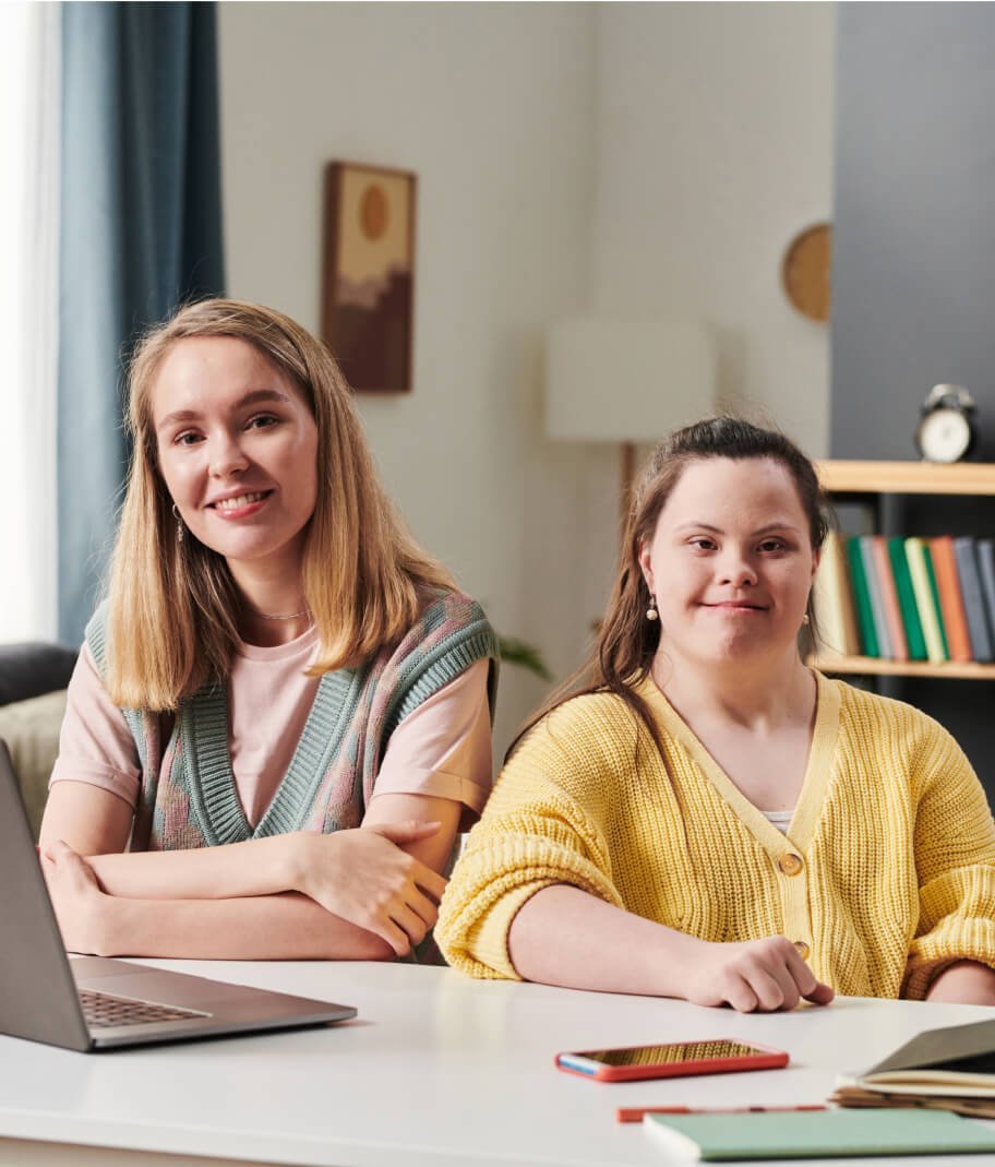 Two girls sitting at a table with a laptop in front of them.