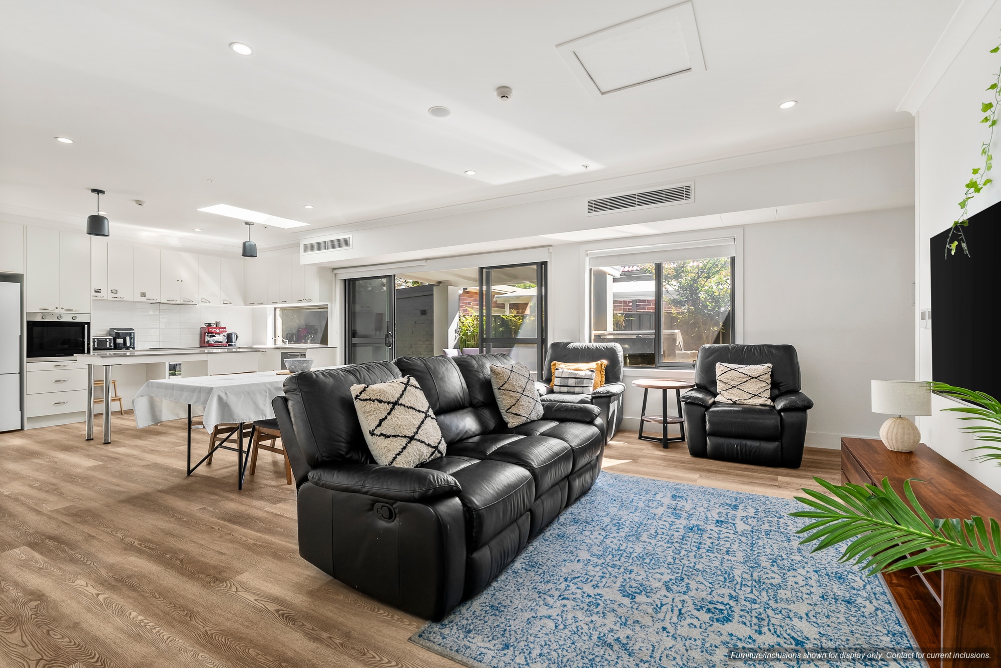 Open plan living and dining area in an Edenbridge Living SDA home, with black leather recliner lounges, a dining table, a kitchen along one wall and sliding doors to a courtyard.