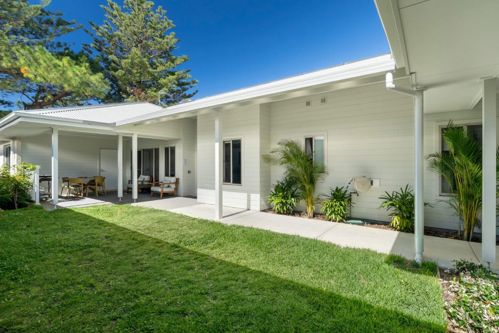 Single-story house with white siding, covered patio, outdoor seating, green lawn, and potted plants under a clear blue sky.