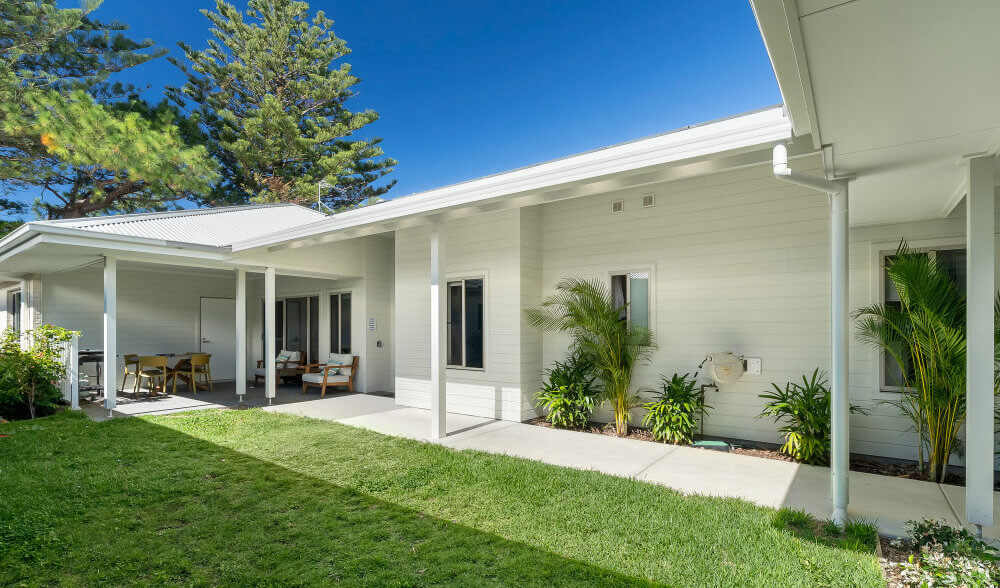 Modern single-story house with white exterior, covered patio, outdoor seating, green lawn, and potted plants on a sunny day.