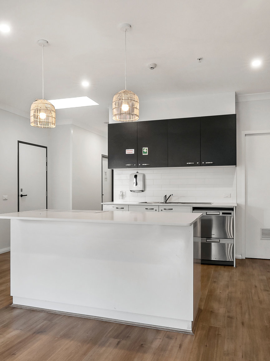 Modern kitchen with black upper cabinets, white island, wood flooring, pendant lights, and stainless steel appliances. White tiled backsplash and minimal decor.