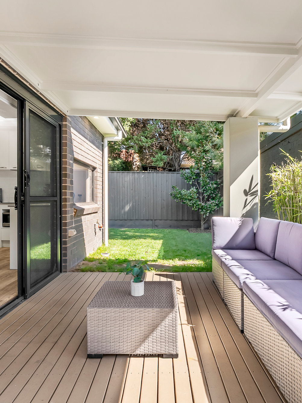 Covered patio with light wood decking, a wicker coffee table with a small plant, a lavender outdoor sofa, and a grassy backyard enclosed by a fence.