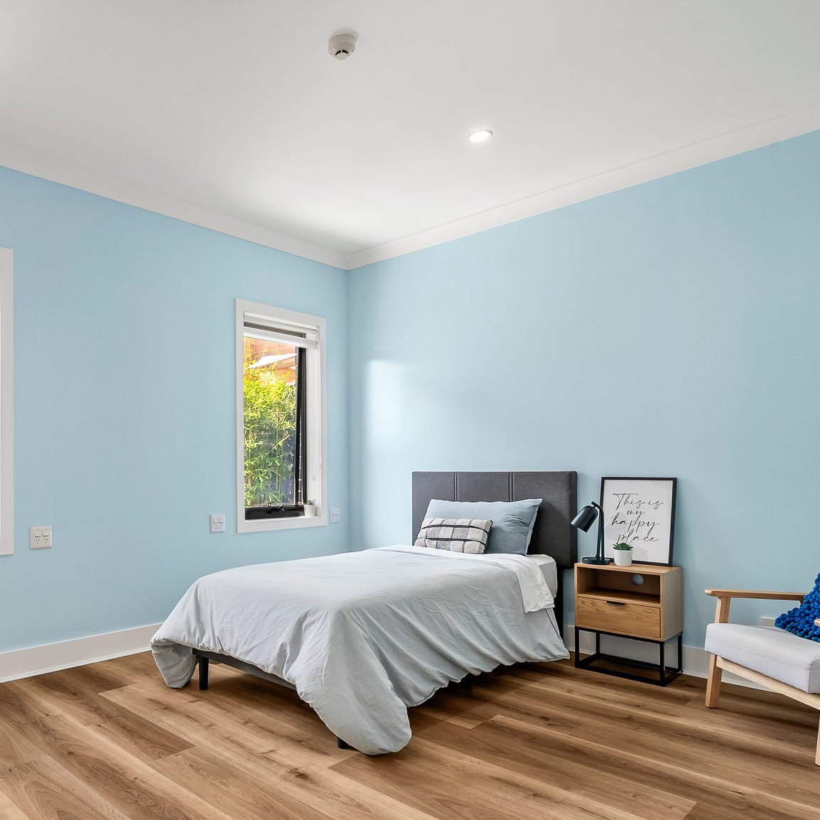 A small bedroom with light blue walls, a single bed with gray bedding, a nightstand with decor, a framed quote, and a window letting in natural light.