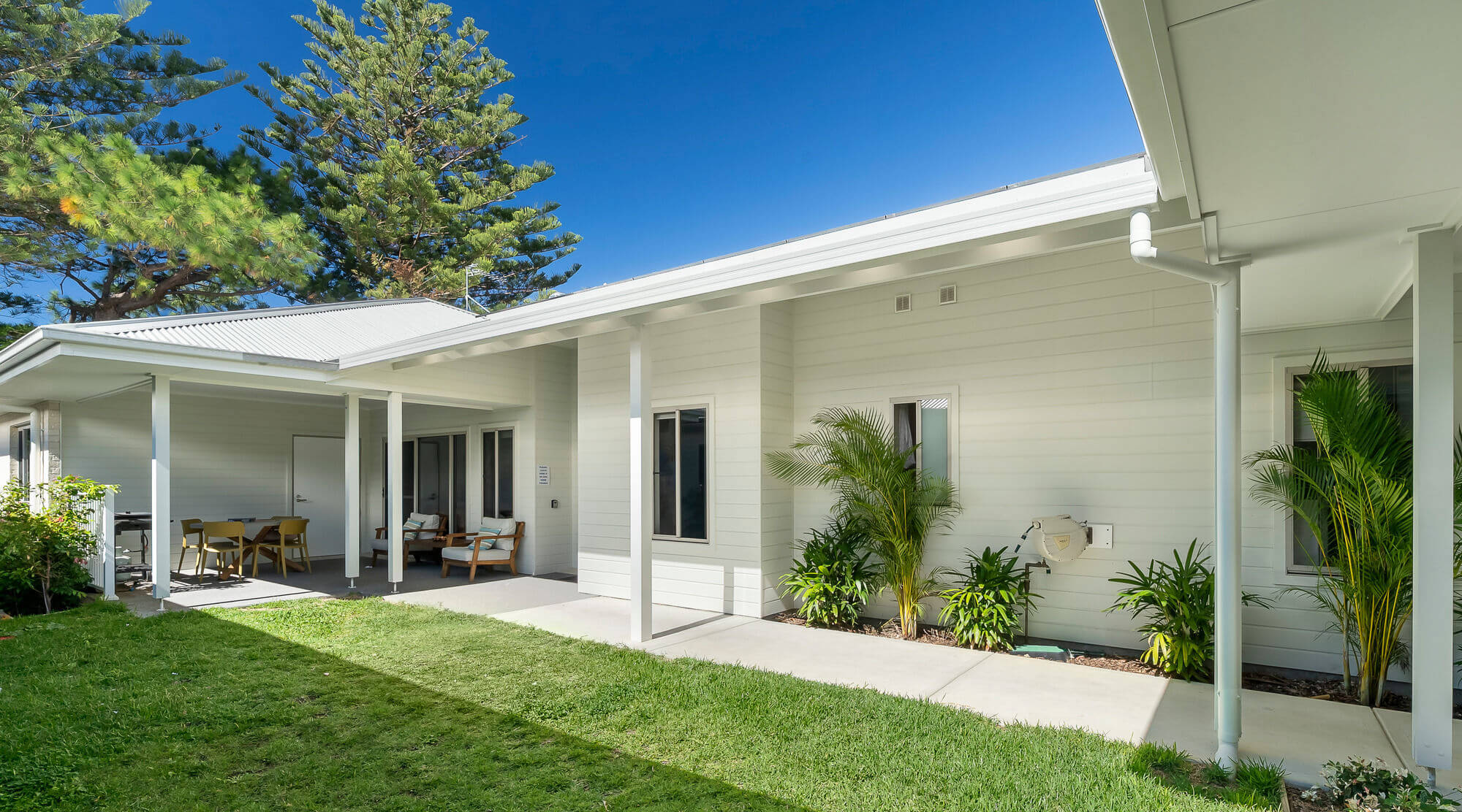 Single-story modern house with light-colored siding, a covered patio area with outdoor seating, green lawn, and potted plants along the exterior wall under a clear blue sky.
