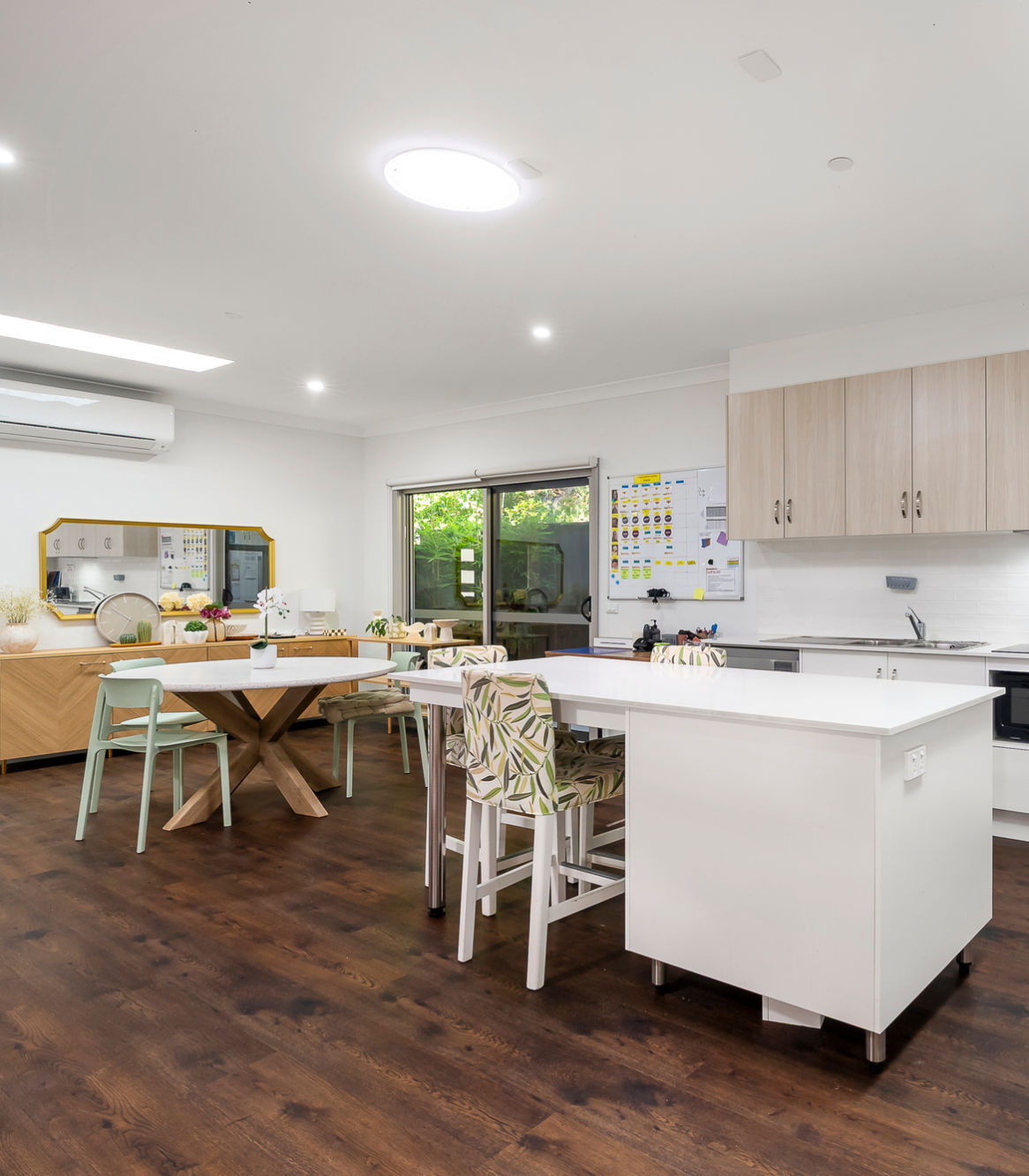 Modern kitchen and dining area with white countertops, wooden cabinets, patterned chairs, large window, and dark wood flooring. A dining table and wall mirror are visible.