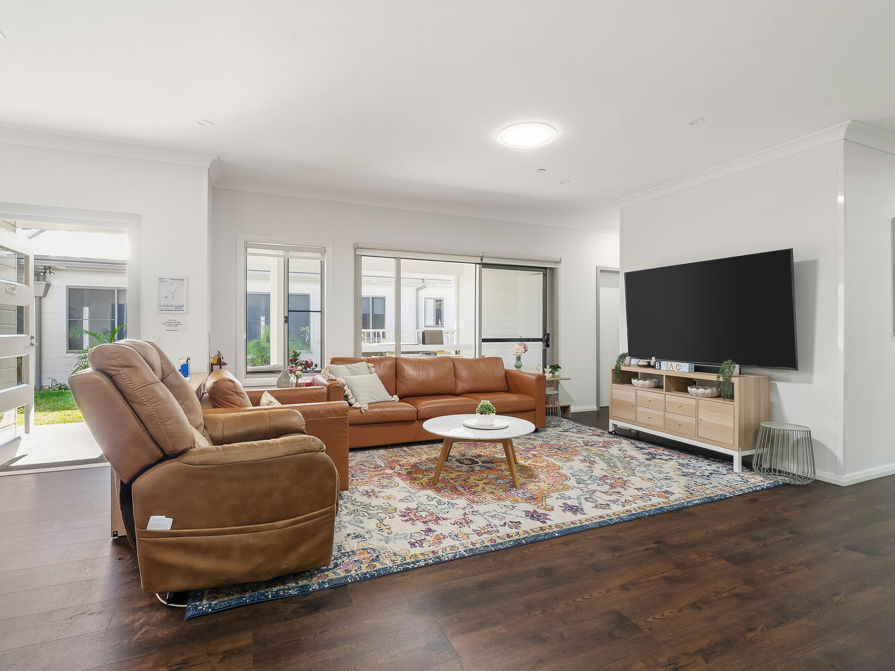 Modern living room with a brown leather sectional, recliner, round coffee table, large TV, wooden console, and a colorful rug on dark wood flooring. Large windows let in natural light.
