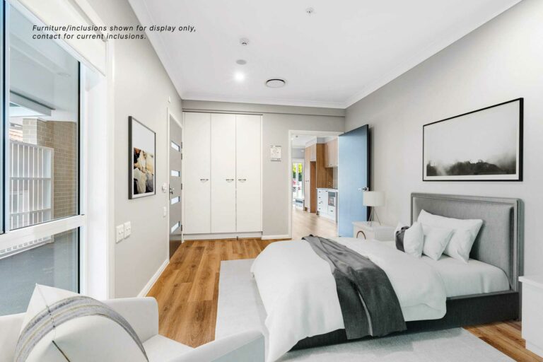 A modern bedroom in Pagewood with a bed, nightstand, chair, and wardrobe. A kitchen area is visible through an open sliding door. Text on the image mentions that the furniture is for display purposes only. This stylish space reflects the contemporary living in Inner South Sydney's 9B district.