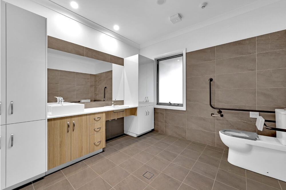 Spacious modern bathroom in Concord, Inner West Sydney, featuring brown wall tiles, white fixtures, a large mirror, accessible toilet with grab bars, and a window.