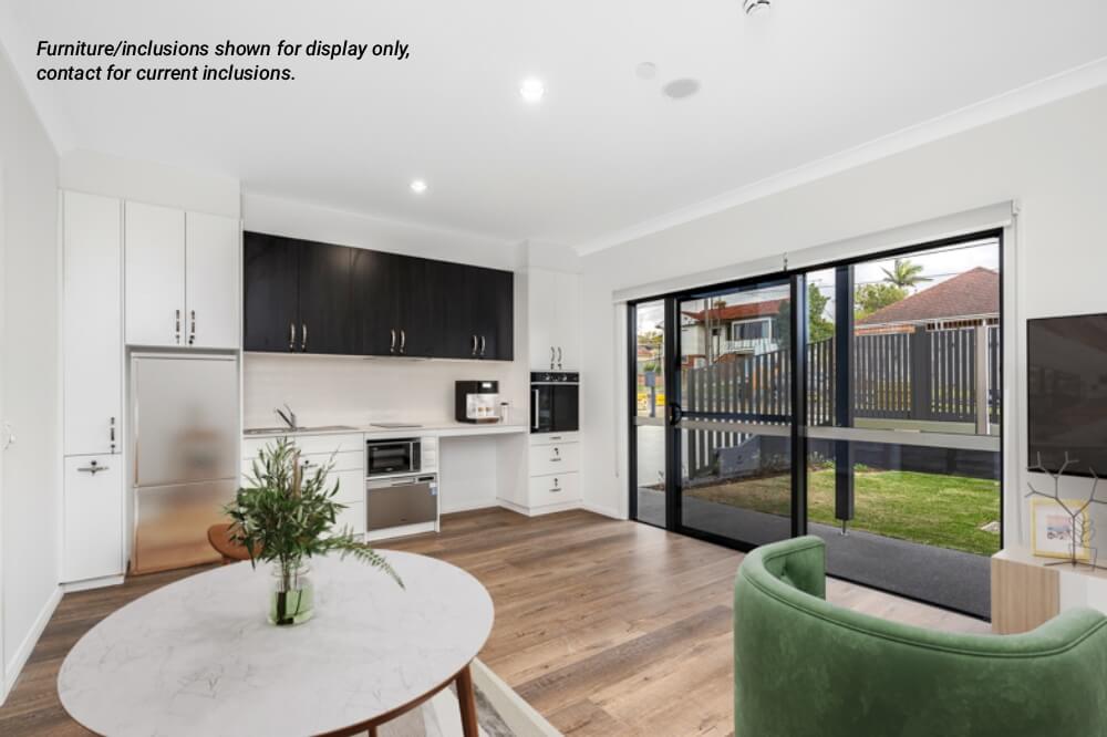 Modern open-plan kitchen and living area in Narraweena with white and dark cabinetry, round wooden dining table, green chair, sliding glass doors, and view of a fenced yard—perfect for enjoying the relaxed Northern Beaches lifestyle.