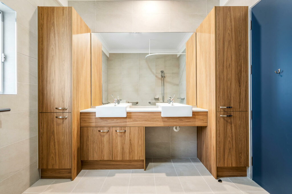 A modern bathroom in the heart of Inner South Sydney, featuring a large mirror, two sinks, wooden cabinets, beige tiles, and a blue door.