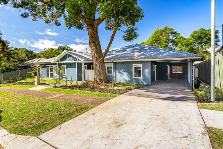 Charming single-story blue house with white trim in Sutherland Shire, featuring a large tree in the front yard, driveway, carport, and landscaped lawn under Sydney’s clear blue sky.