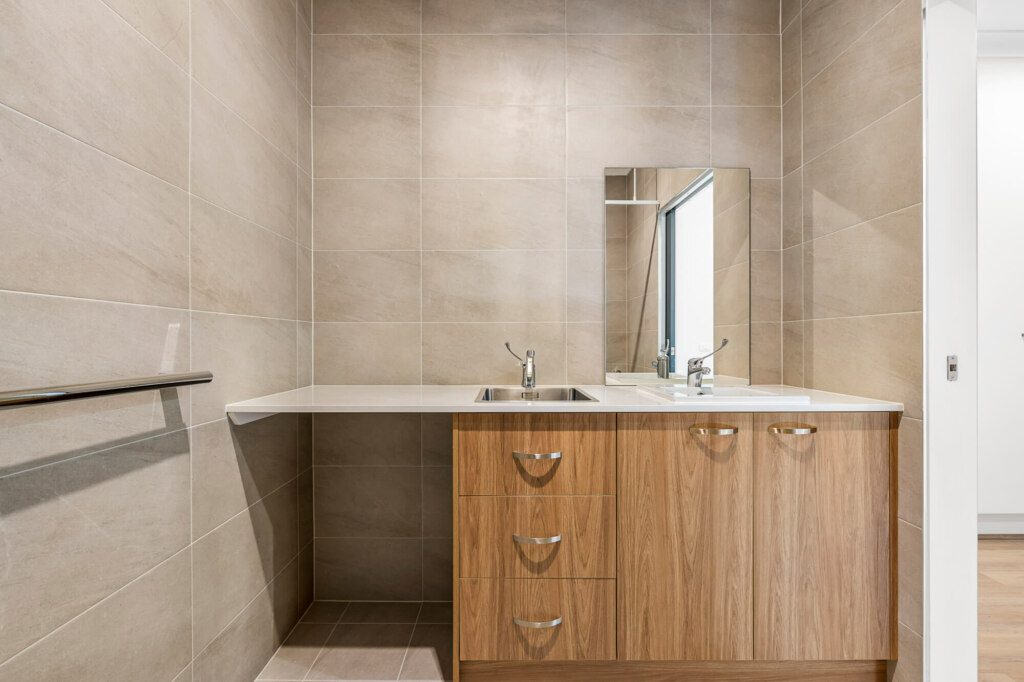 A compact bathroom in the Sutherland Shire, Sydney, featuring beige tiled walls, a wooden vanity with a countertop, two sinks, mirrors, and storage drawers underneath. The sleek design of 30A brings both functionality and style to this serene space.