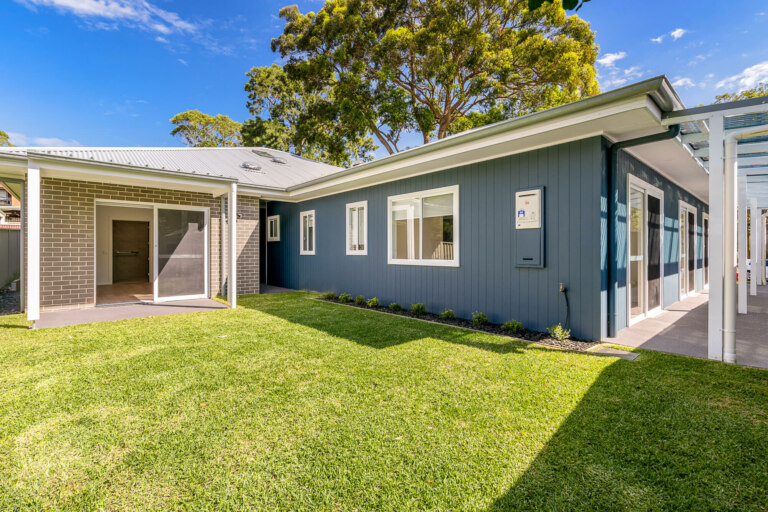 A modern single-story home in the heart of Sutherland Shire, Sydney, boasts a light gray exterior, a well-maintained lawn, large windows, and a covered patio area under a clear blue sky. 