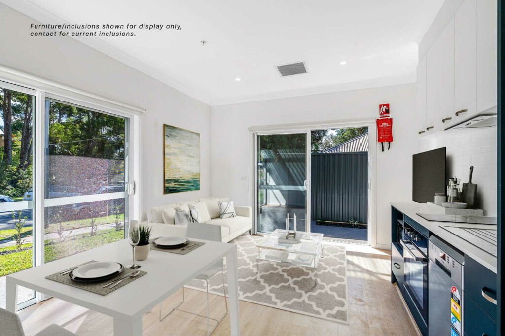Modern, bright living area with light wood floors, white walls, and large windows offering garden views. This Sutherland Shire gem is furnished with a sectional sofa, a small dining table set for two, and a compact kitchen area.